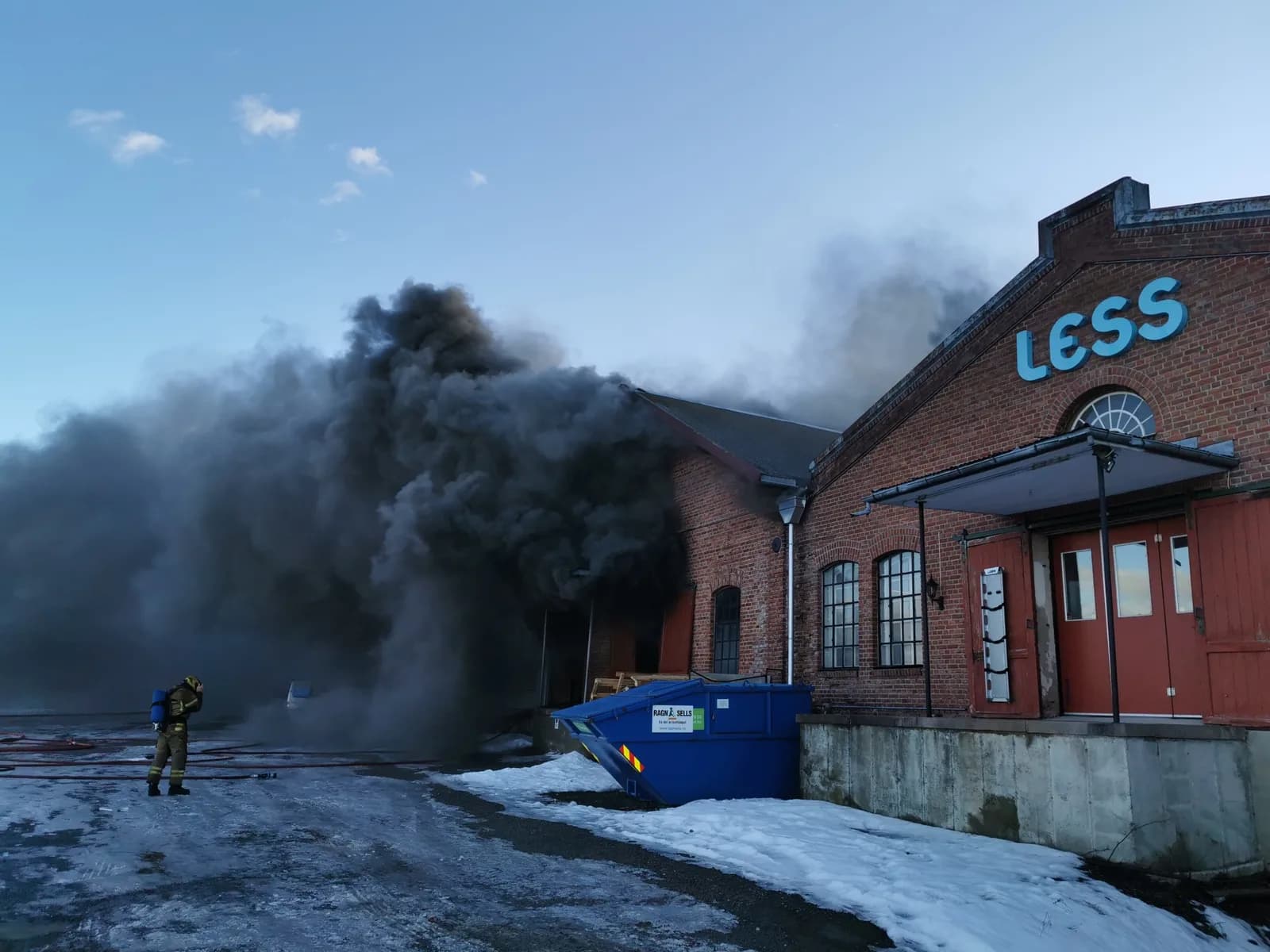 15:43. Black smoke pours from the roof of the building with the LESS sign. A single firefighter on the scene, snow on the ground.