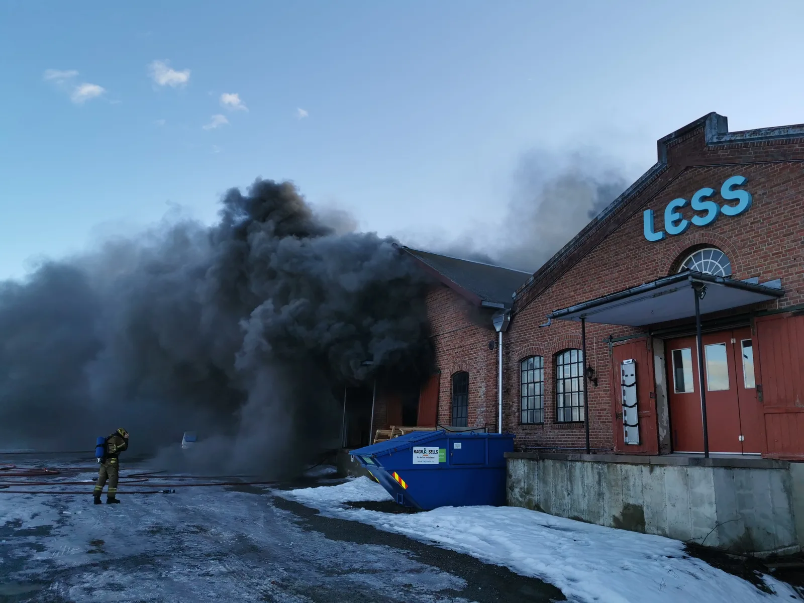 15:43. Svart røyk velter ut av taket på lokalene med LESS-skiltet. Én brannmann alene på plassen, snø på bakken.
